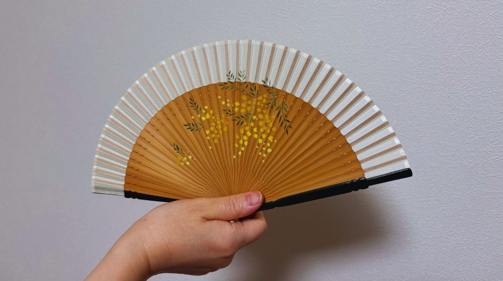 ミモザ柄の扇子を広げてあおぐ様子。やわらかな風を感じる日常のひととき